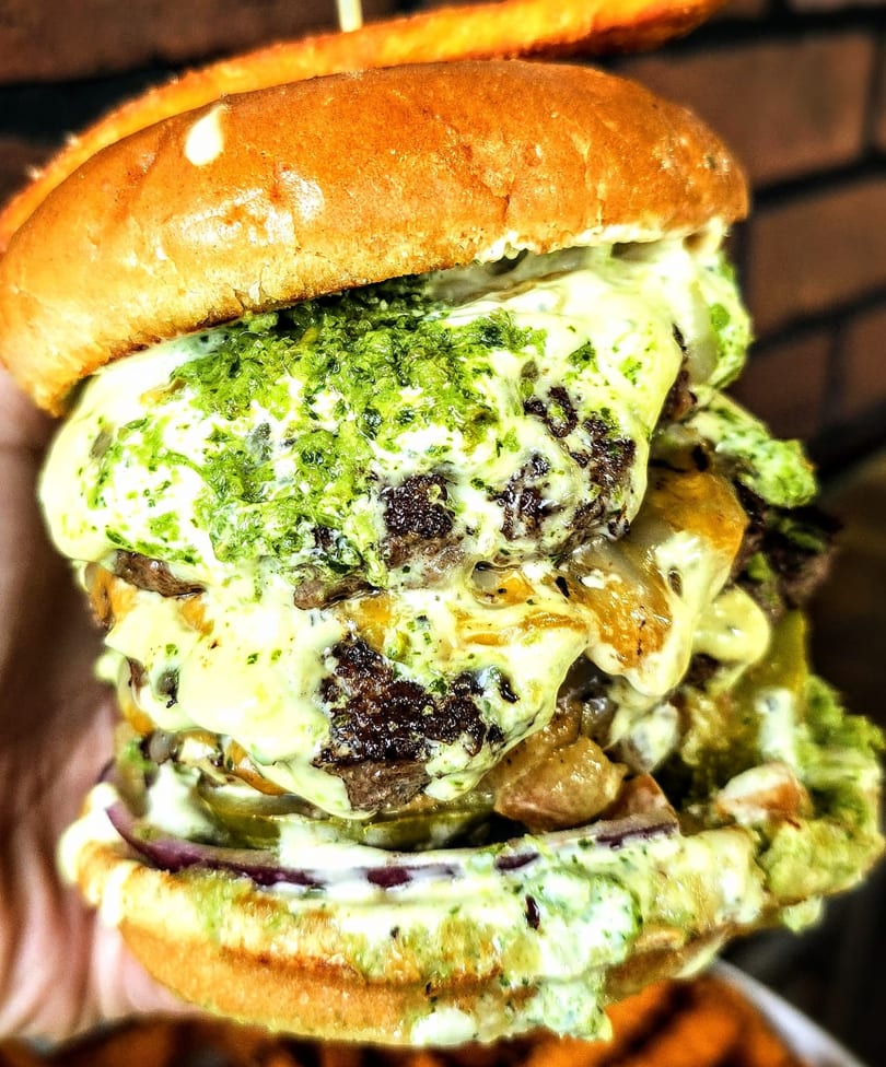 A towering Burger Xpress burger with multiple patties, melted cheese, and green relish.